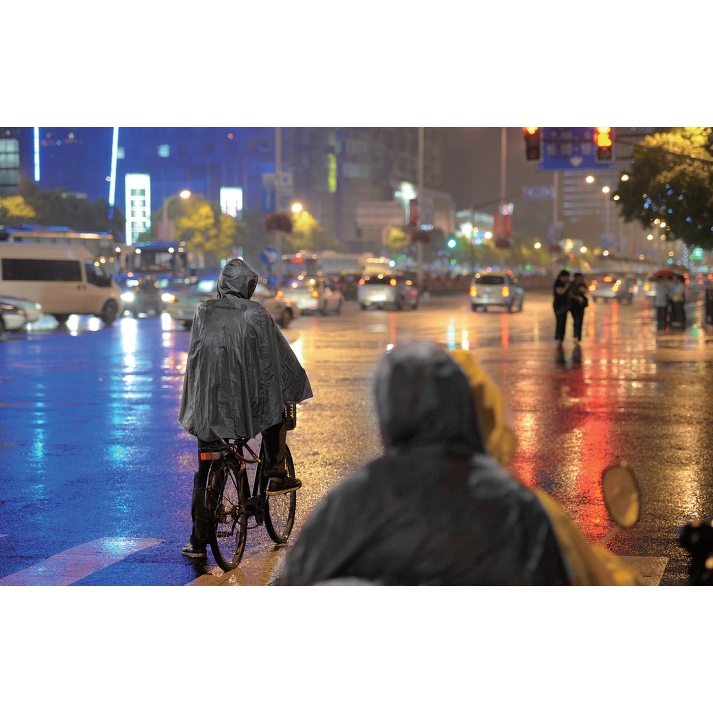 Person mit schwarzem Regenponcho fährt bei Regen auf einem Fahrrad durch eine beleuchtete Stadtstraße mit reflektierendem Boden.