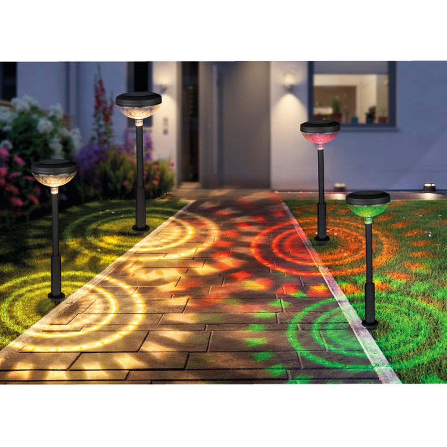 Vier Solarlampen entlang eines Gartenwegs bei Nacht, jede Lampe wirft bunte, kreisförmige Lichtmuster in Gelb, Rot oder Grün auf den Boden; moderne Hausfassade im Hintergrund.