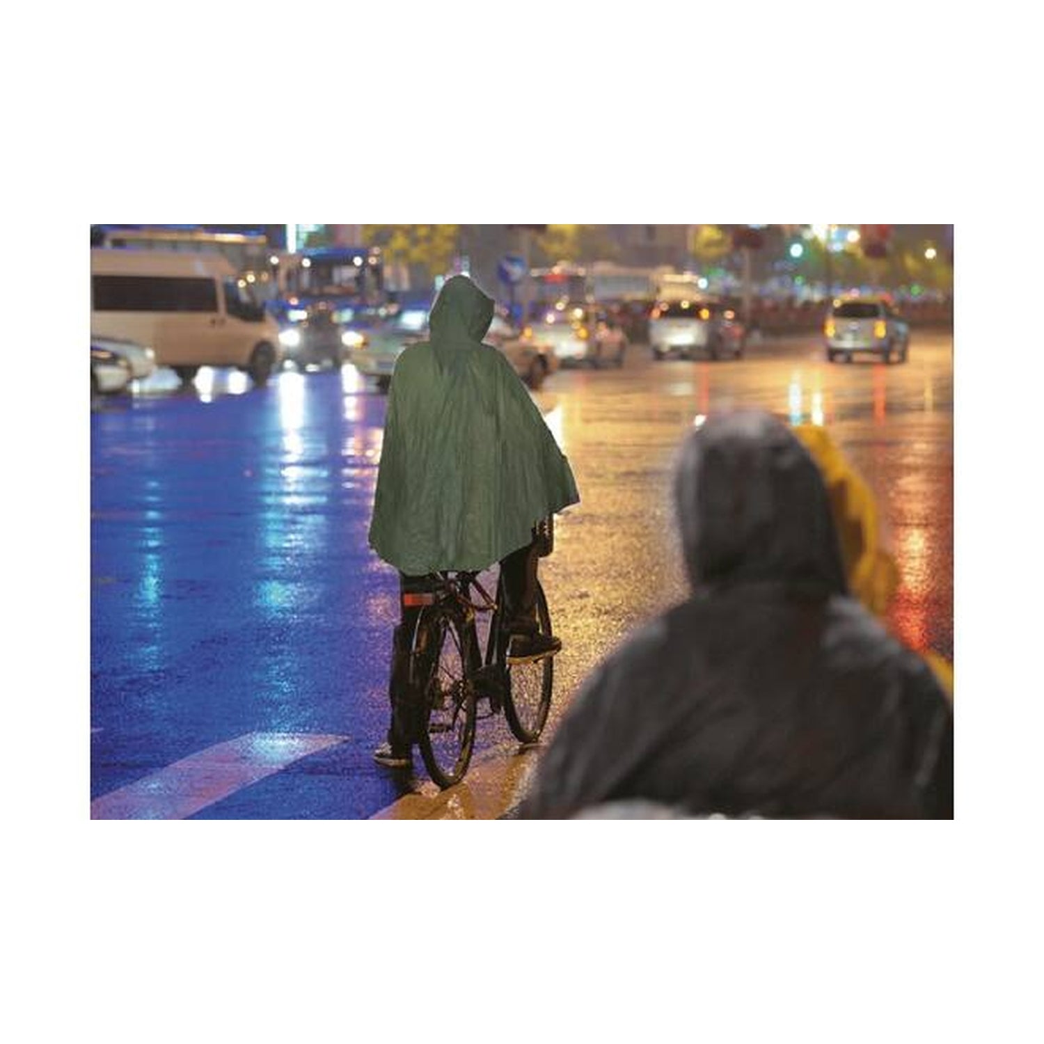 Person mit grünem Regenponcho fährt bei Regen auf einem Fahrrad durch eine nasse, beleuchtete Stadtstraße.