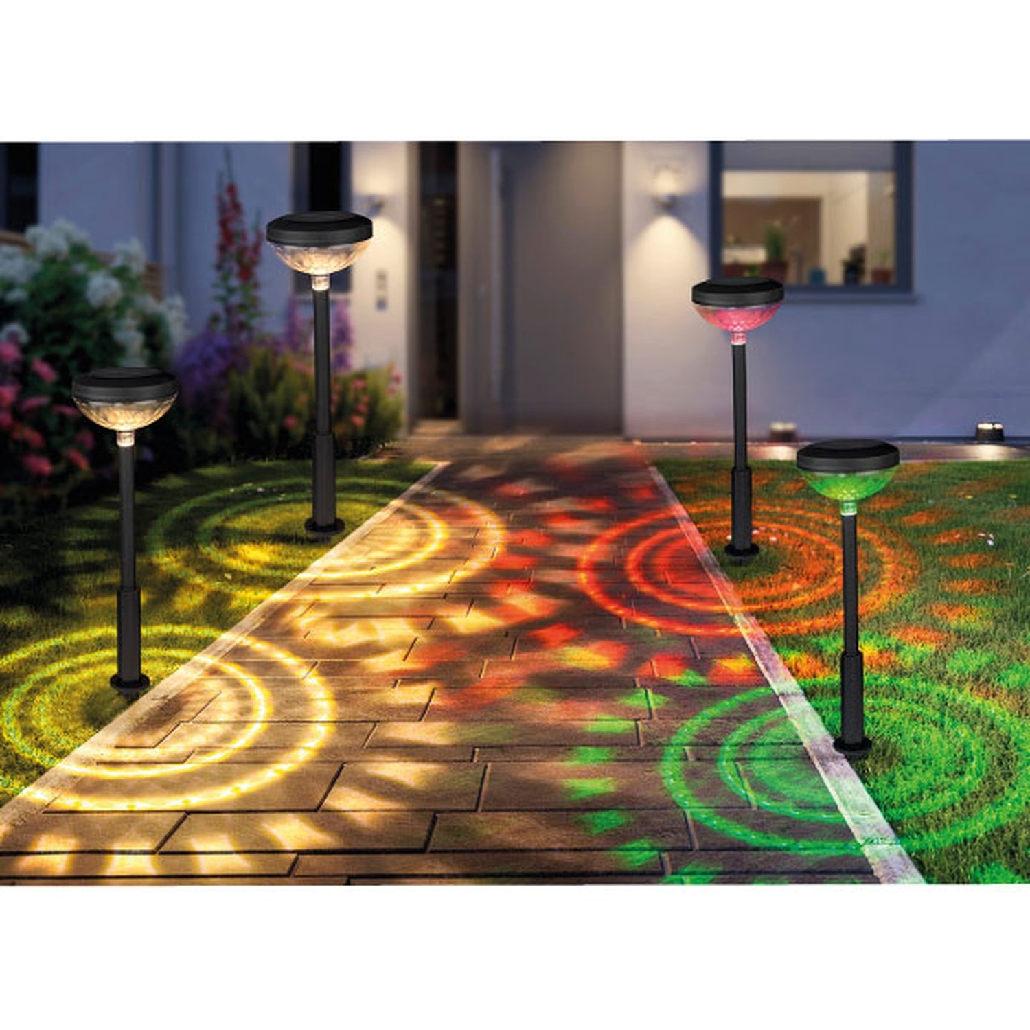 Vier Solarlampen entlang eines Gartenwegs bei Nacht, jede Lampe wirft bunte, kreisförmige Lichtmuster in Gelb, Rot oder Grün auf den Boden; moderne Hausfassade im Hintergrund.