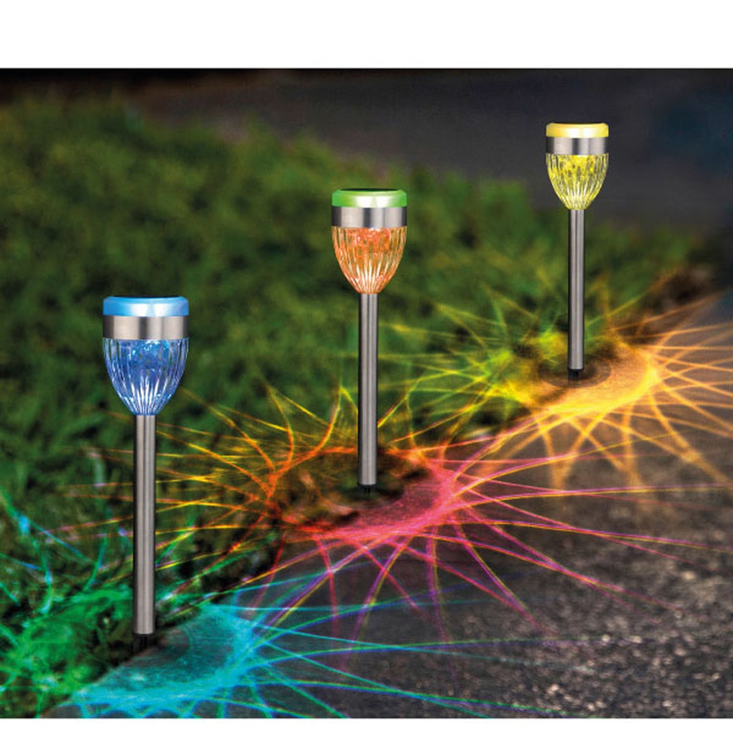 Drei solarbetriebene Gartenleuchten aus Edelstahl mit unterschiedlich farbigen Leuchtköpfen in Blau, Orange und Gelb erzeugen bei Dunkelheit leuchtende, sternförmige Muster auf Rasen und Weg.