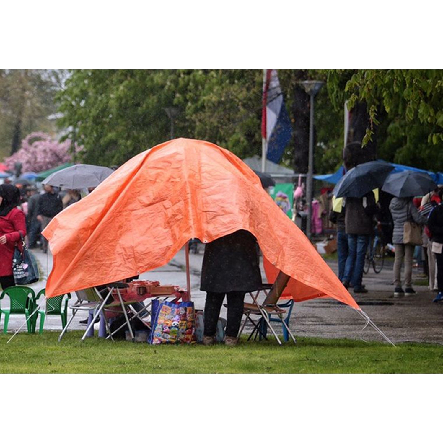 Orangefarbene Plane als provisorisches Zeltdach auf einer Wiese gespannt, darunter Tische und Stühle mit mehreren Personen bei Regen.