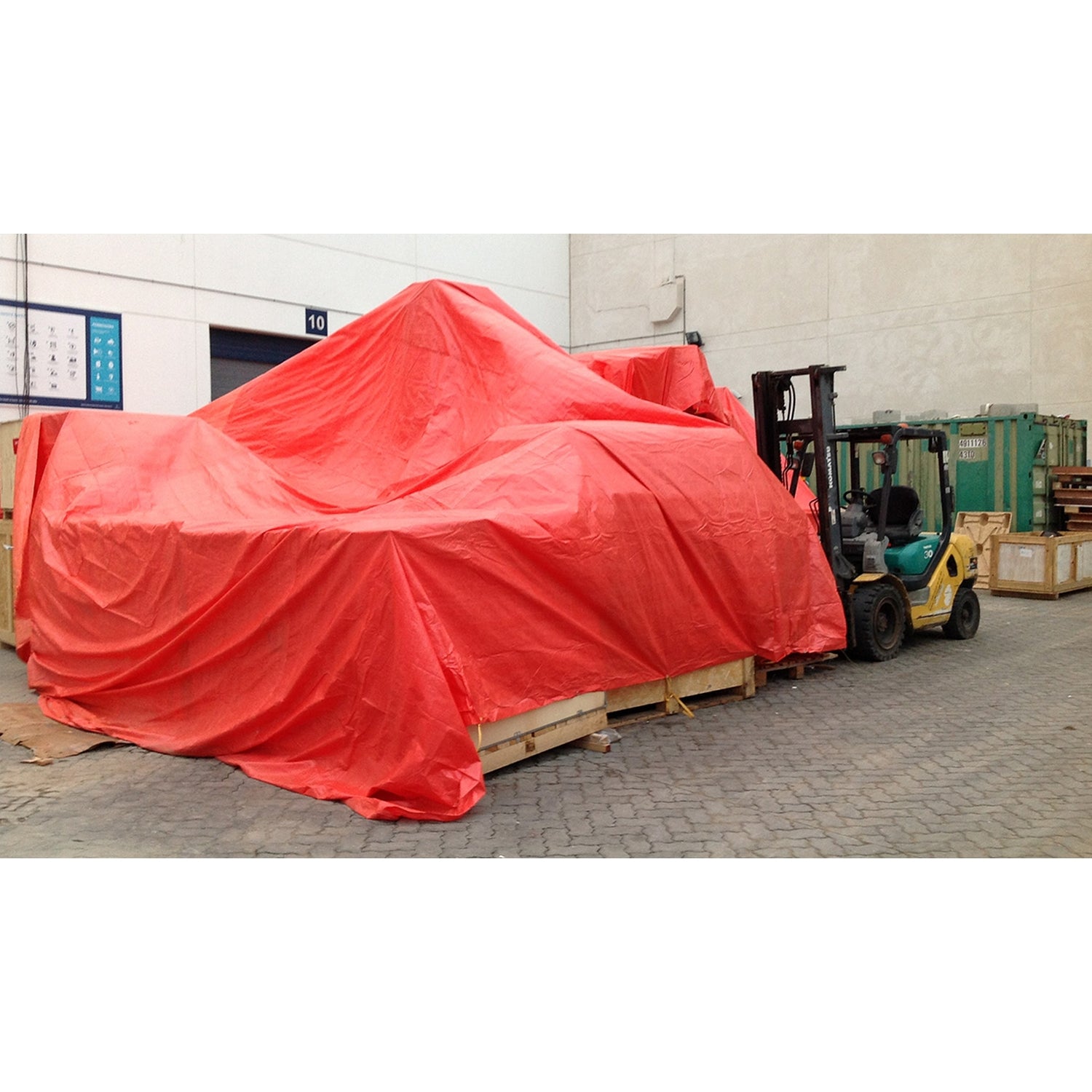 Große, mit einer orangefarbene Gewebeplane abgedeckte Gegenstände auf Holzkisten in einem Industriehof; rechts steht ein Gabelstapler.