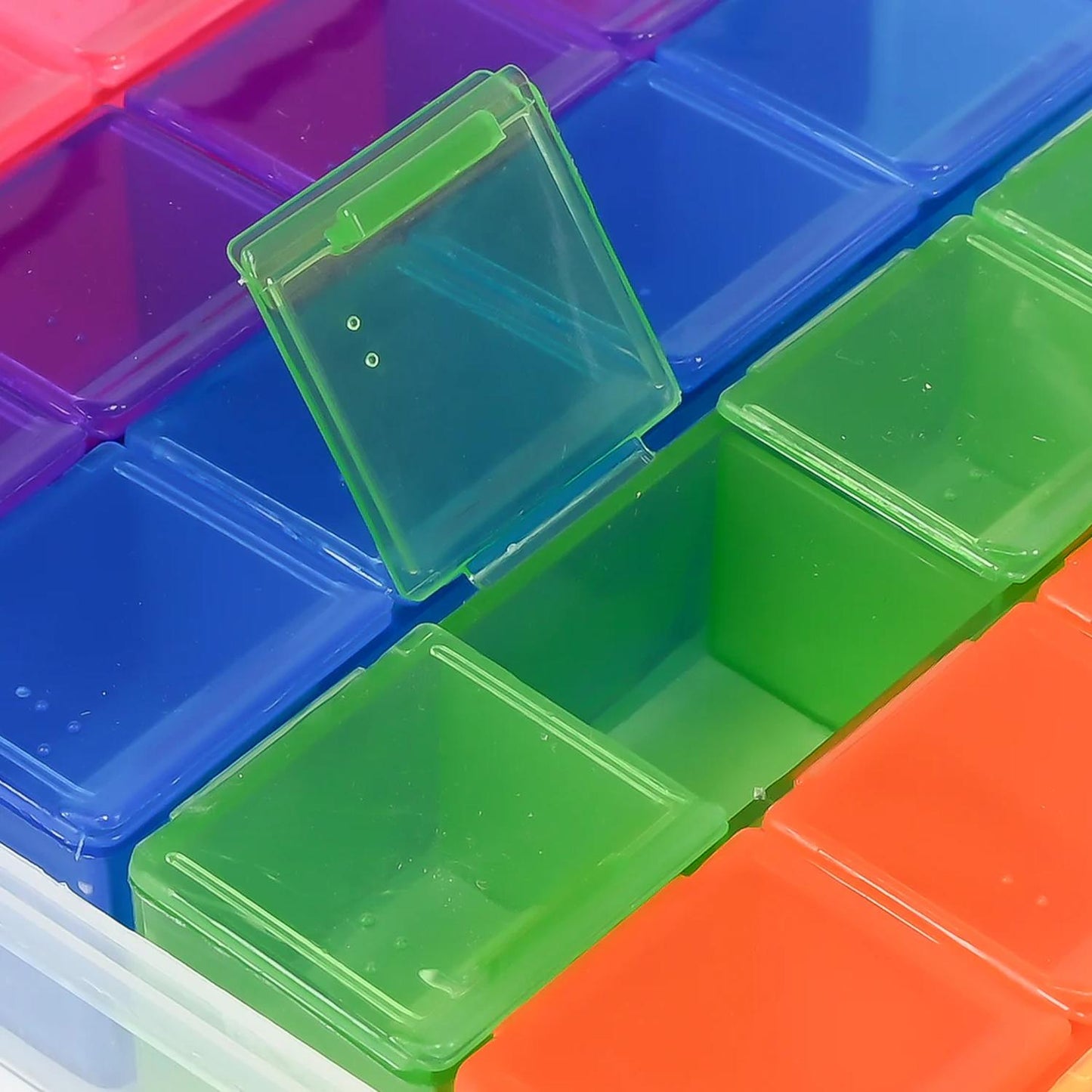 Detailaufnahme einer farbigen Pillenbox mit geöffnetem grünem Fach, umgeben von blauen, violetten und orangefarbenen Fächern, transparenter Kunststoff sichtbar.